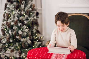 Yeşil sandalyede oturan ve Noel zamanı sırasında bilgisayar tablet ile oynayan sevimli çocuk portresi. Tatil mevsim.