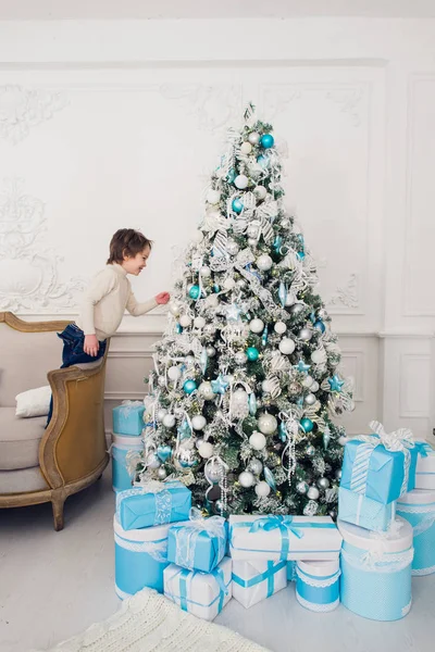 Divertido niño de pie en un sofá cerca del árbol de Navidad con luces de colores y mirando a su reflejo en la bola de Navidad —  Fotos de Stock