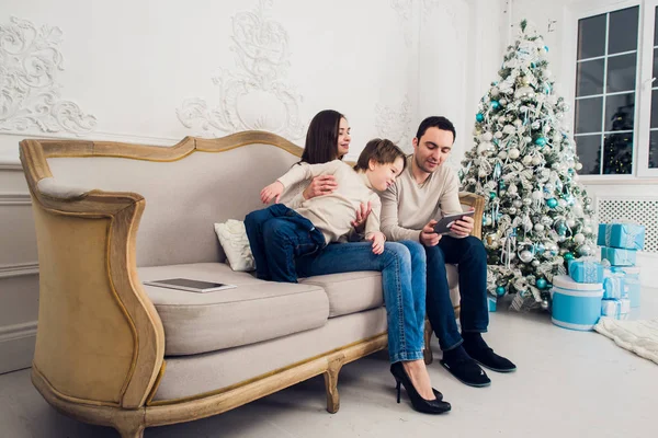 Mañana de Navidad, familia alegre sentada en la sala de estar divirtiéndose con la tableta digital que Santa Claus le trajo, detrás del árbol decorado — Foto de Stock