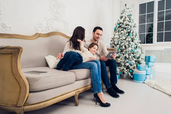 Alegre familia sentada en la sala de estar divirtiéndose con la tableta digital que Santa Claus le trajo, detrás del árbol de navidad decorado —  Fotos de Stock