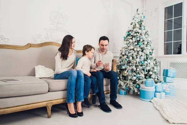 Alegre familia sentada en la sala de estar divirtiéndose con la tableta digital que Santa Claus le trajo, detrás del árbol de navidad decorado — Foto de Stock
