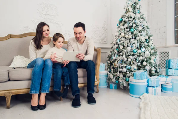 Alegre familia sentada en la sala de estar divirtiéndose con la tableta digital que Santa Claus le trajo, detrás del árbol de navidad decorado — Foto de Stock