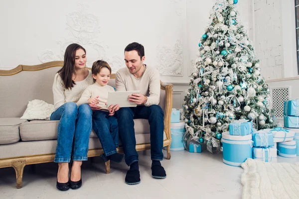 Alegre familia sentada en la sala de estar divirtiéndose con la tableta digital que Santa Claus le trajo, detrás del árbol de navidad decorado —  Fotos de Stock