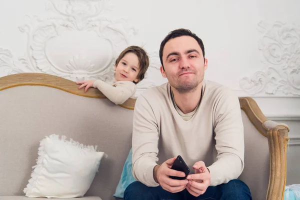 累了沮丧的父亲坐在沙发上，使用手机和儿子想要和他一起玩 — 图库照片