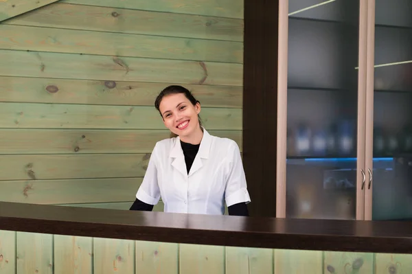 Mujer en la recepción sonríe y da la bienvenida al visitante virtual en el spa o clínica moderna. Documentos con acuerdo en su mano . — Foto de Stock