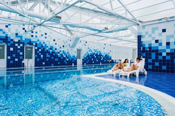 Duas mulheres bonitas estão deitadas na piscina de chaise-longue dentro de casa e conversando . — Fotografia de Stock