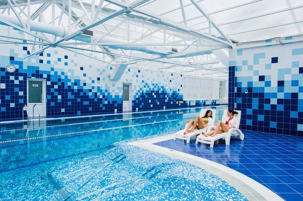 Dos hermosas mujeres están tumbadas en la chaise-longue junto a la piscina en el interior y hablando . —  Fotos de Stock