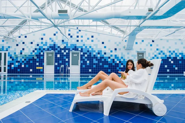 Dos hermosas mujeres están tumbadas en la chaise-longue junto a la piscina en el interior y hablando . —  Fotos de Stock