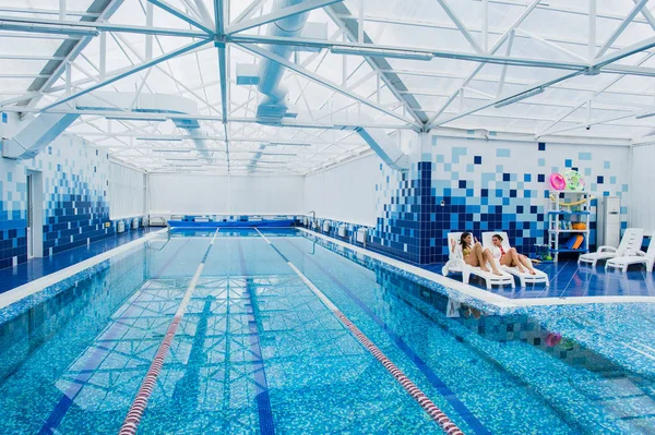 Duas mulheres bonitas estão deitadas na piscina de chaise-longue dentro de casa e conversando . — Fotografia de Stock