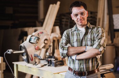 Koruyucu gözlük yakışıklı marangoz kameraya bakarak ve atölyede ahşap onun nesne yakın dururken gülümseyen