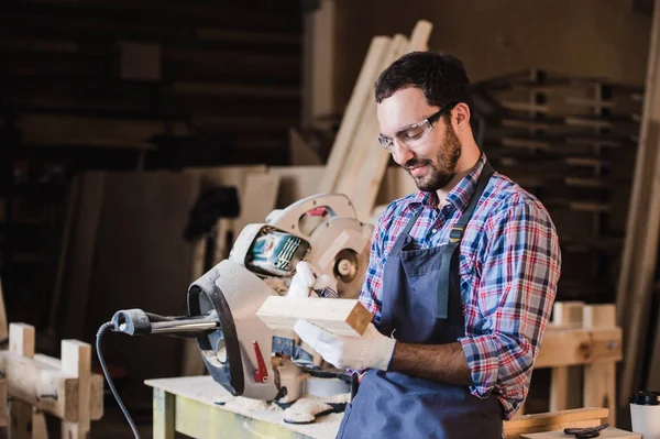 Hermoso carpintero en gafas protectoras está mirando a la cámara y sonriendo mientras está de pie cerca de su objeto de madera en el taller —  Fotos de Stock