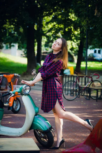 Moda miejska. Plenerowej dość młoda kobieta ubrana w kratkę sukienka stojący w pobliżu skuter w parku na zewnątrz — Zdjęcie stockowe