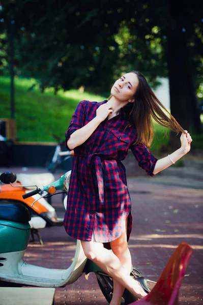 Mujer de moda en vestido a cuadros posando y tirando de su pelo cerca de scooter sobre fondo del parque de verano — Foto de Stock