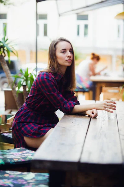 Por encima de la vista mujer bonita en vestido a cuadros violeta sentado en la mesa en la terraza en la cafetería. Ella sostiene un vaso de agua, mirando a la cámara — Foto de Stock