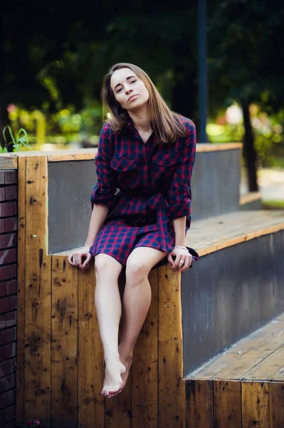 Flicka klädd i rutig klänning sitter i skatepark tittar på kameran. — Stockfoto