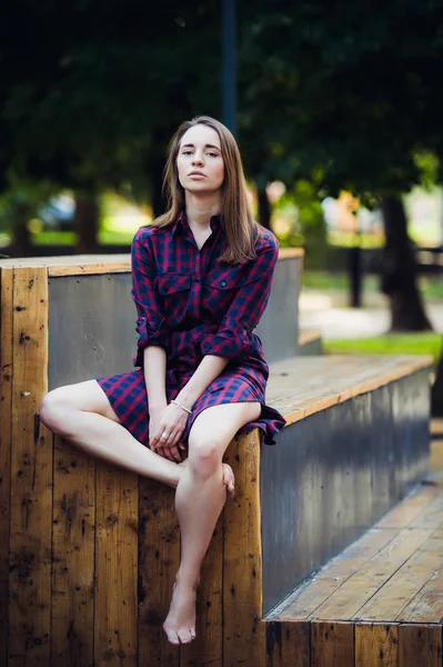 Flicka klädd i rutig klänning sitter i skatepark tittar på kameran. — Stockfoto