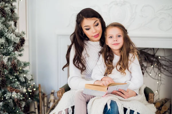 Joyeux Noël et Joyeuses Fêtes, Jolie jeune maman lisant un livre à sa jolie fille près de l'arbre à l'intérieur . — Photo