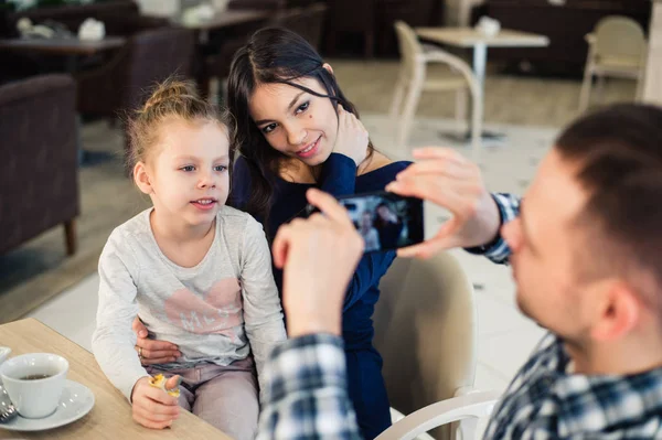 Familia, paternidad, tecnología, concepto de personas - padre feliz tomando fotos de su pequeña hija y esposa por teléfono inteligente cenando en el restaurante — Foto de Stock