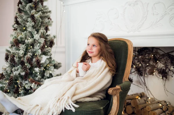 クリスマス ツリーと暖炉の家で牛乳を飲む休日寝具で毛布に包まれたアームチェアに座っているパジャマを身に着けている女の子 — ストック写真
