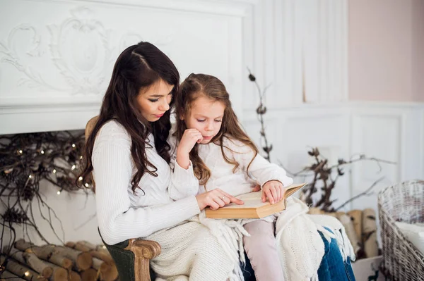 Feliz Navidad y Felices Fiestas, bastante joven mamá leyendo un libro a su linda hija cerca del árbol en el interior . — Foto de Stock