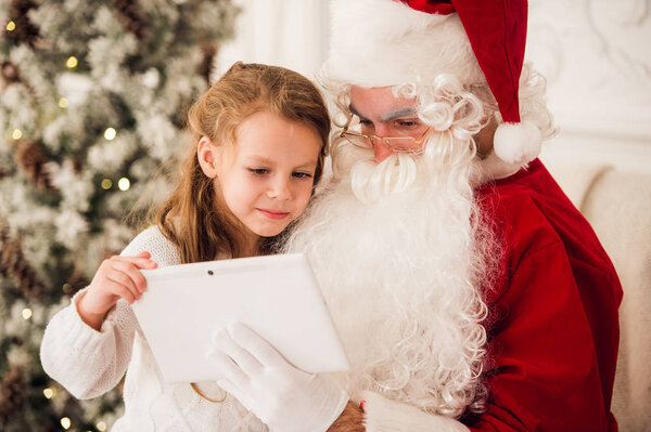 Little girl sitting with Santa using tablet on the armchair at home living room