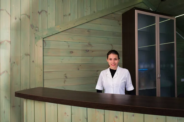 Mujer en la recepción sonríe y da la bienvenida al visitante virtual en el spa o clínica moderna. Documentos con acuerdo en su mano . — Foto de Stock