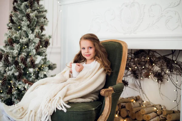 Het dragen van pyjama zittend op fauteuil gewikkeld in een deken met vakantie beddengoed door kerstboom en open haard consumptiemelk thuis meisje — Stockfoto