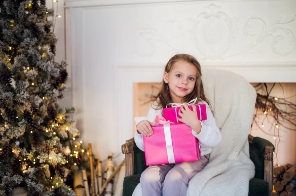 Mooi meisje in de buurt van kerstboom uitpakken presenteert zittend op een stoel — Stockfoto