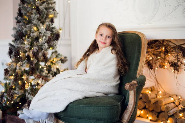 Schattig klein meisje zit door de kerstboom, gewikkeld in een warme deken thuis — Stockfoto