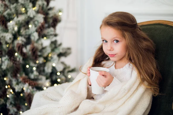 Petite fille portant un pyjama assis sur un fauteuil enveloppé dans une couverture avec literie de vacances près de l'arbre de Noël et cheminée boire du lait à la maison — Photo