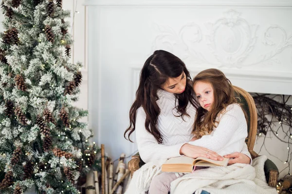 Joyeux Noël et Joyeuses Fêtes, Jolie jeune maman lisant un livre à sa jolie fille près de l'arbre à l'intérieur . — Photo