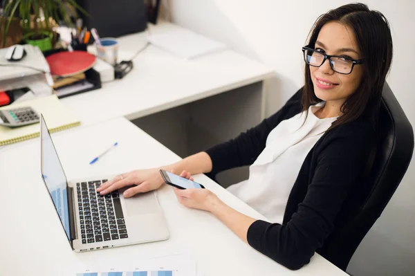 Buena charla de negocios. Joven alegre hermosa mujer en gafas hablando en el teléfono móvil y el uso de la computadora portátil con sonrisa mientras está sentada en su lugar de trabajo —  Fotos de Stock