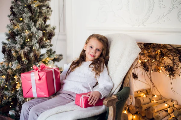 Gelukkig kind meisje zittend op fauteuil bedekt met een deken tegen ingerichte kerst open haard — Stockfoto