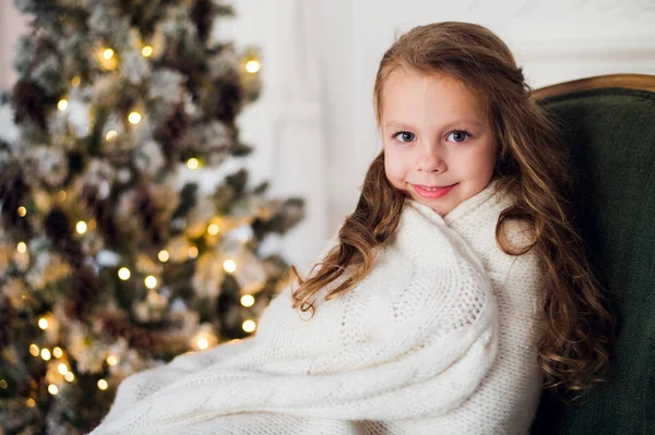 Schattig klein meisje zit door de kerstboom, gewikkeld in een warme deken thuis — Stockfoto