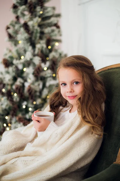 Het dragen van pyjama zittend op fauteuil gewikkeld in een deken met vakantie beddengoed door kerstboom en open haard consumptiemelk thuis meisje — Stockfoto