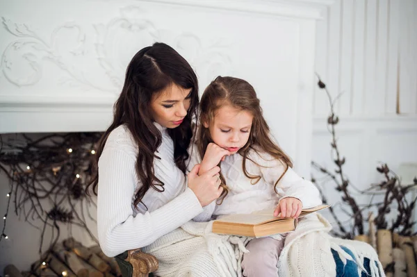 Feliz Navidad y Felices Fiestas, bastante joven mamá leyendo un libro a su linda hija cerca del árbol en el interior . — Foto de Stock