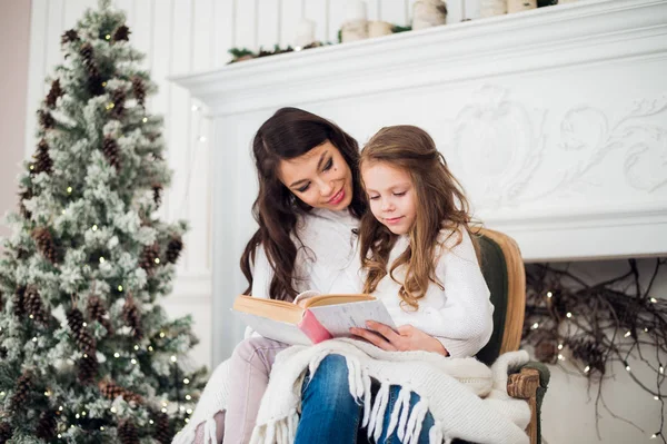 Feliz Navidad y Felices Fiestas, bastante joven mamá leyendo un libro a su linda hija cerca del árbol en el interior . — Foto de Stock