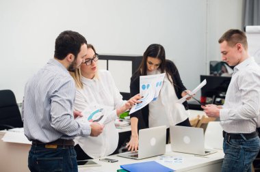 Four young business people working as a team gathered around laptop computer in an open plan modern office clipart