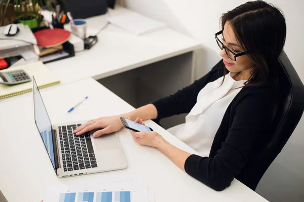 Buena charla de negocios. Joven alegre hermosa mujer en gafas hablando en el teléfono móvil y el uso de la computadora portátil con sonrisa mientras está sentada en su lugar de trabajo —  Fotos de Stock