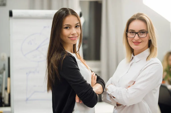 Dos compañeras de trabajo de negocios de pie en una oficina y sonriendo positivamente a la cámara mientras sostienen la carpeta de papeleo — Foto de Stock