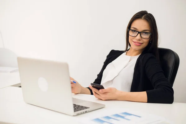 Boa conversa de negócios. Jovem mulher bonita alegre em óculos falando no telefone celular e usando laptop com sorriso enquanto está sentado em seu local de trabalho — Fotografia de Stock