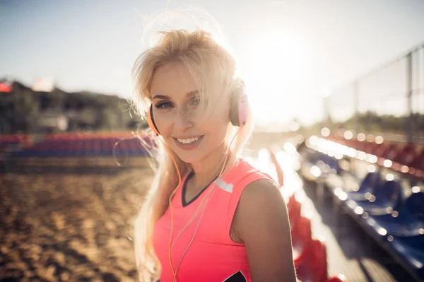 Sport buiten foto van mooie jonge blonde vrouw in roze kleurrijke pak, luisteren naar muziek op de hoofdtelefoon aan het strand — Stockfoto