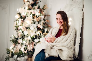 Ev holding fincan sıcak kahve sıcak örme kazak giyiyor oturma ile genç güzel kadın. Noel ağacı süsleme ve ışıklar odasında