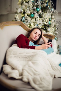 Genç kadın o liyng kanepe önünde evde Noel ağacı olarak cep telefonuna metin mesajlarını kontrol.