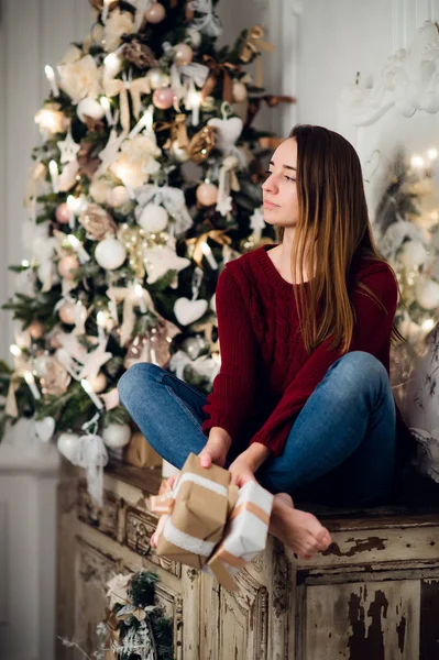 Жінка з різдвяним подарунком, що сидить на старовинному комоді, прикрашеному Різдвом. Ялинка на фоні — стокове фото