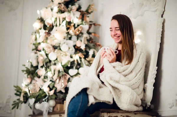温かみのあるニットのセーターを着ているホット コーヒーのホーム開催カップを座っていると若い美しい女性。クリスマス ツリーの装飾、部屋の照明 — ストック写真