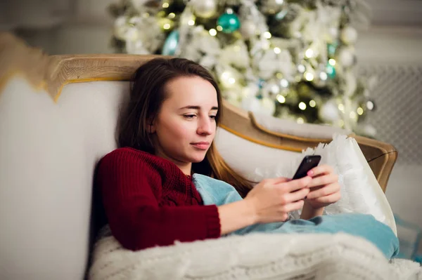 Mujer joven mirando sus mensajes de texto en el teléfono móvil mientras liyng sofá en frente del árbol de Navidad en casa . — Foto de Stock