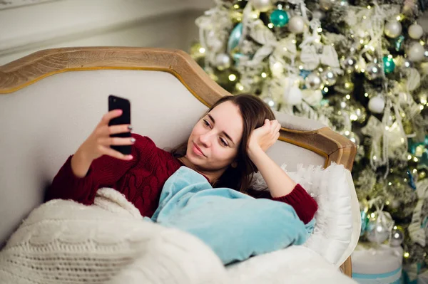 Mujer joven con teléfono móvil en casa tumbada en un sofá frente al abeto — Foto de Stock