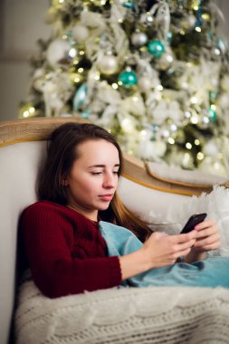 Genç kadın o liyng kanepe önünde evde Noel ağacı olarak cep telefonuna metin mesajlarını kontrol.