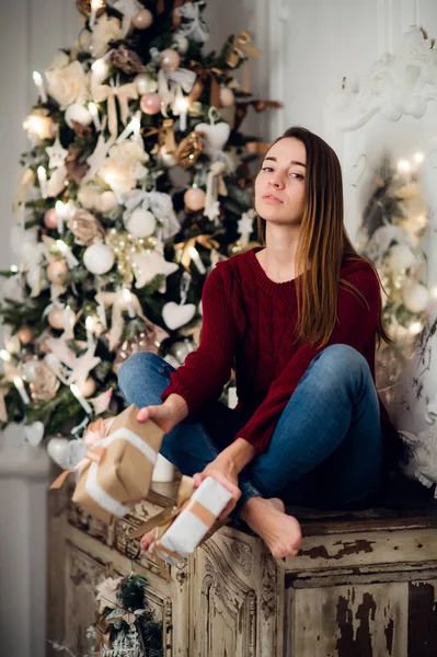 Woman with a Christmas Gift sitting on acncient commode decorated for xmas. Fir tree at background — Stock Photo, Image